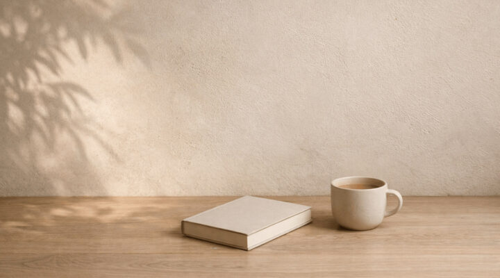 Libro cerrado y taza de cerámica sobre una mesa de madera clara junto a una pared beige iluminada por luz natural con sombras suaves de hojas.