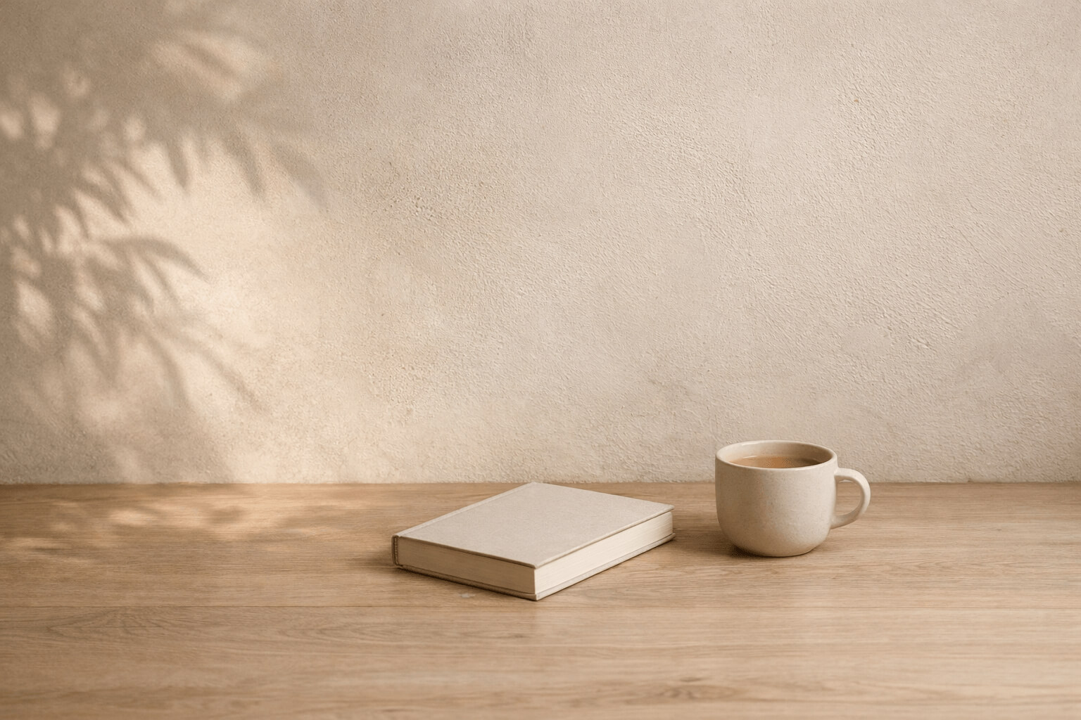 Libro cerrado y taza de cerámica sobre una mesa de madera clara junto a una pared beige iluminada por luz natural con sombras suaves de hojas.