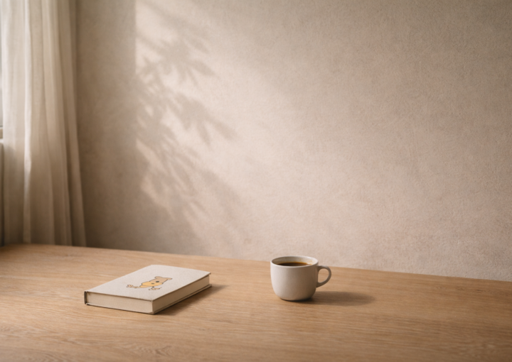 Cuaderno infantil cerrado y taza blanca sobre mesa de madera junto a una ventana con luz natural suave y sombra de hojas en pared beige.