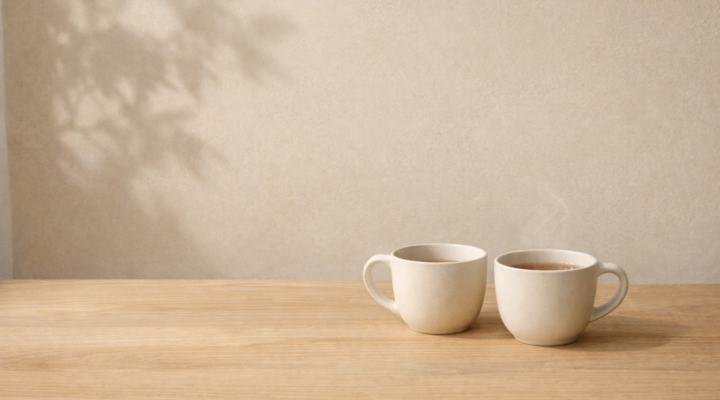Dos tazas de cerámica sobre mesa de madera clara con luz natural lateral y sombra de planta en pared beige.