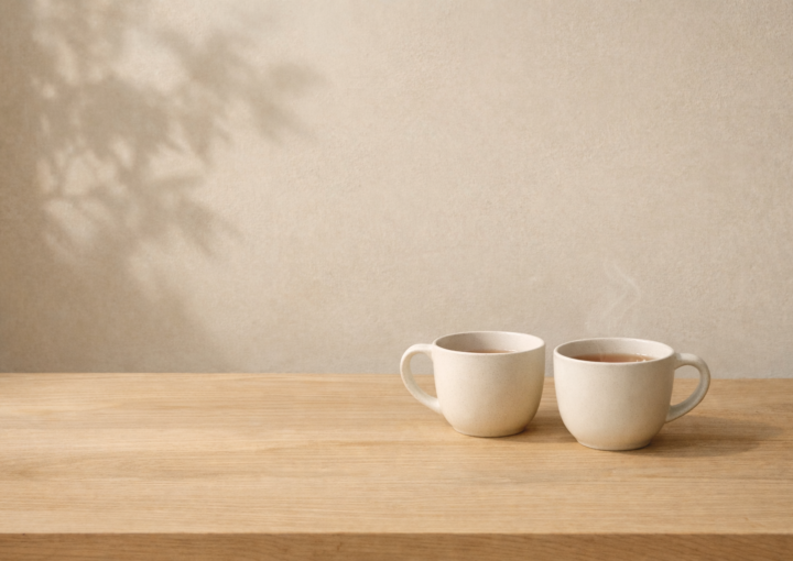 Dos tazas de cerámica sobre mesa de madera clara con luz natural lateral y sombra de planta en pared beige.