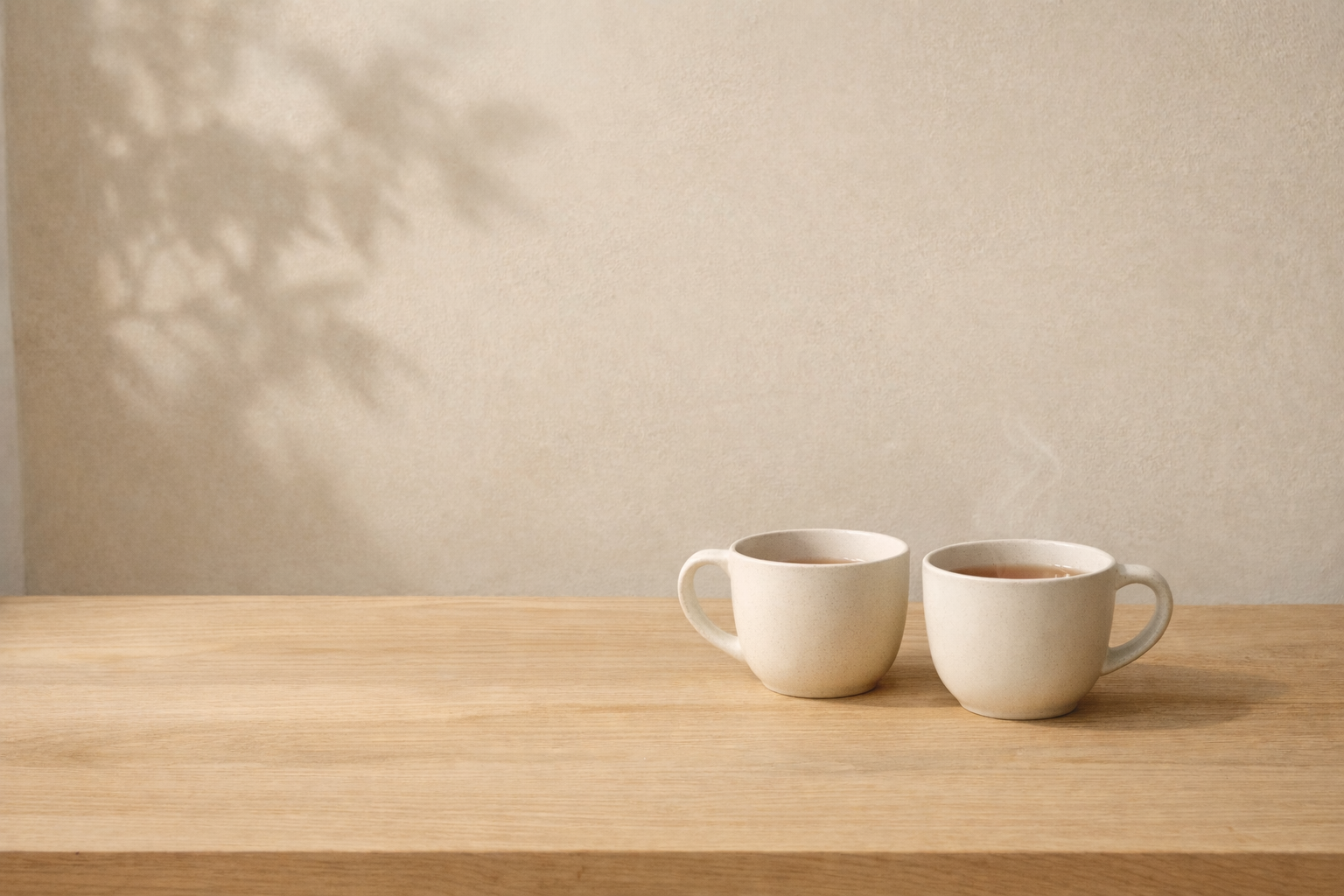 Dos tazas de cerámica sobre mesa de madera clara con luz natural lateral y sombra de planta en pared beige.