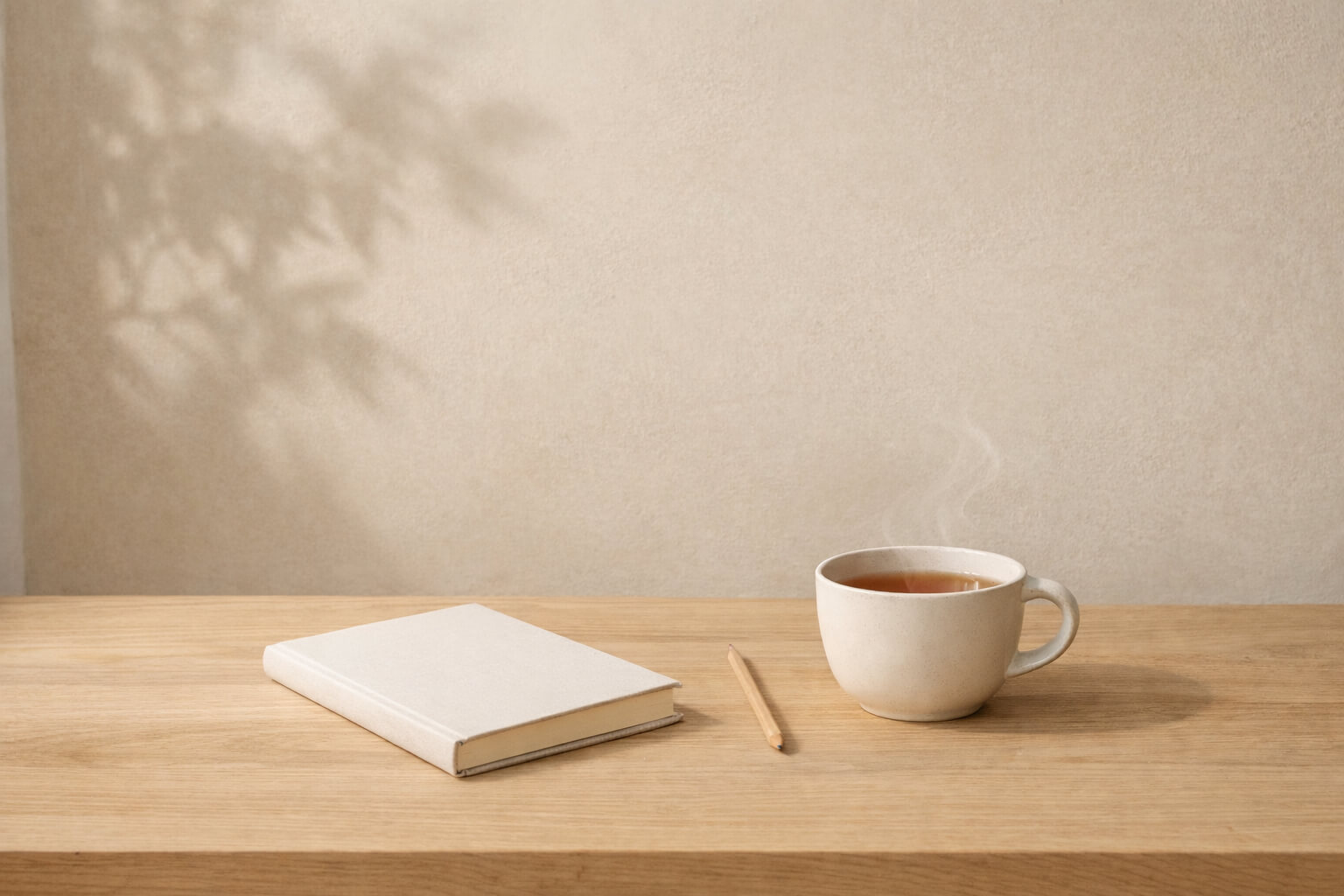 Cuaderno cerrado, lápiz y taza de té sobre mesa de madera clara con luz natural y sombra de planta en pared beige.